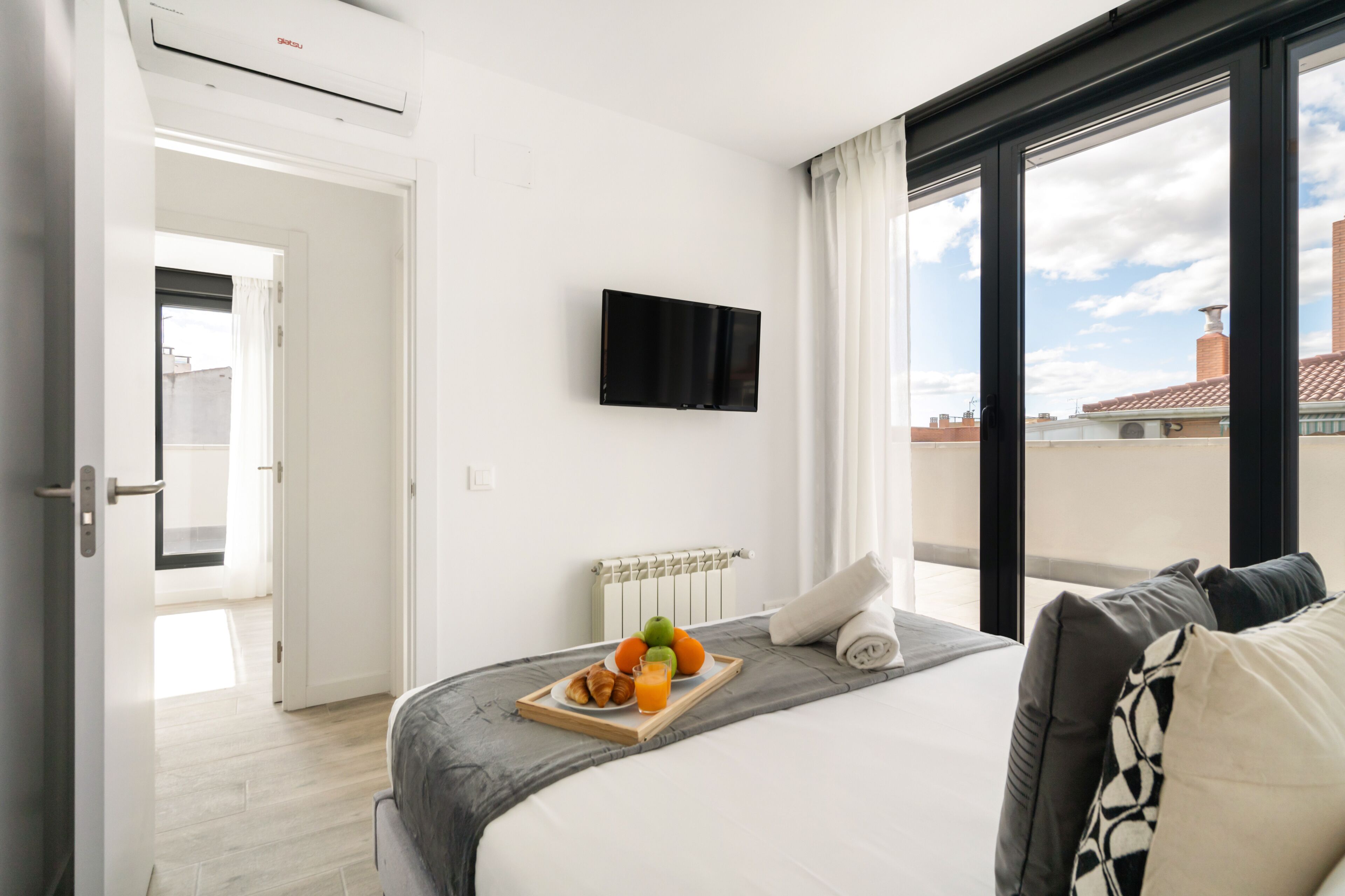 A hotel room with a bed, a flat-screen TV mounted on the wall, a breakfast tray with fruits and pastries, and a view of the city through large windows.