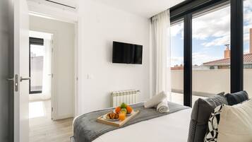 A hotel room with a bed, a flat-screen TV mounted on the wall, a breakfast tray with fruits and pastries, and a view of the city through large windows.