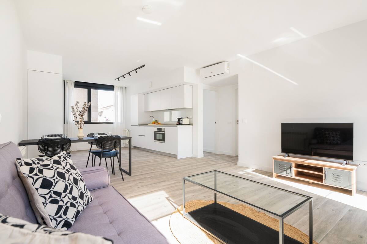 A modern living space with a sofa, a glass coffee table, a TV, and a kitchen area with a dining table and chairs.
