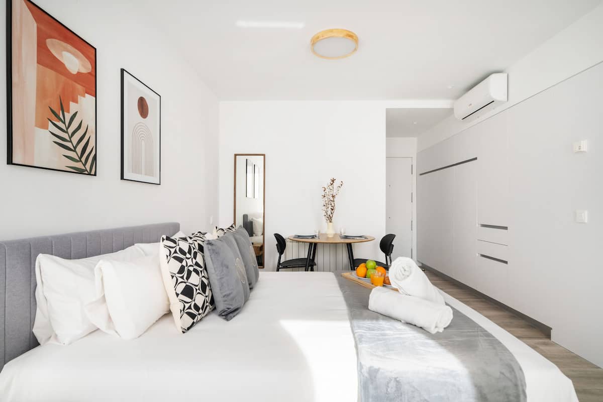 A modern bedroom with a bed, a gray sofa, a dining table with chairs, and a small kitchen area.