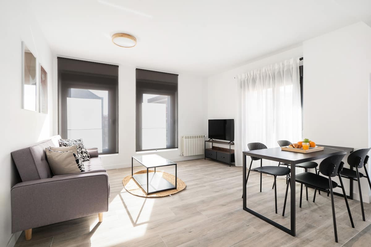 A modern living room with a grey sofa, a glass coffee table, a dining table with chairs, and a TV.