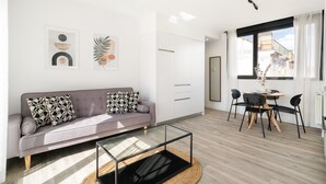 A modern living room with a grey sofa, black and white patterned cushions, a glass coffee table, and a dining area with a round table and chairs.