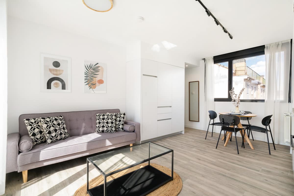 A modern living room with a grey sofa, black and white patterned cushions, a glass coffee table, and a dining area with a round table and chairs.