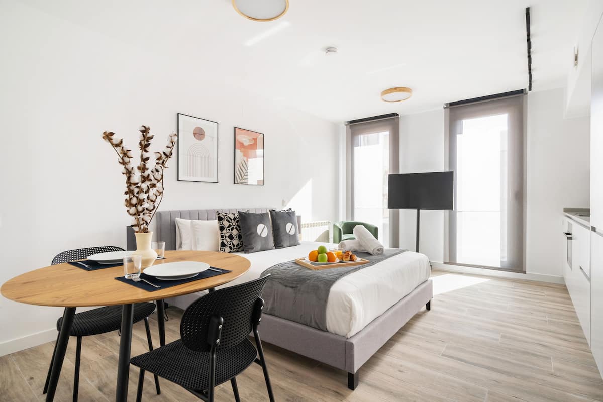 A modern hotel room with a bed, a dining table with chairs, a flat-screen TV, and artwork on the wall.