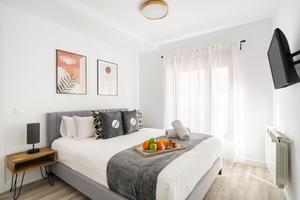 A modern hotel room with a large bed, a bedside table with a lamp, a tray with fruits and pastries, and a TV mounted on the wall.