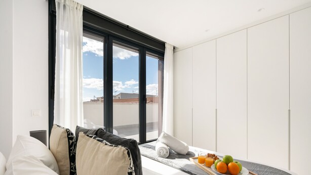 A modern hotel room with a large window, a bed with a gray headboard, and a breakfast tray with fruits and juice.