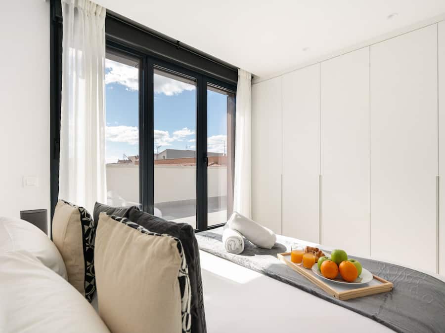 Une chambre d’hôtel moderne dotée d’une grande fenêtre, d’un lit avec une tête de lit grise et d’un plateau de petit-déjeuner avec des fruits et du jus.