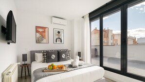 A modern bedroom with a large window, a bed with a tray of fruit and a bottle, a flat-screen TV, and artwork on the wall.