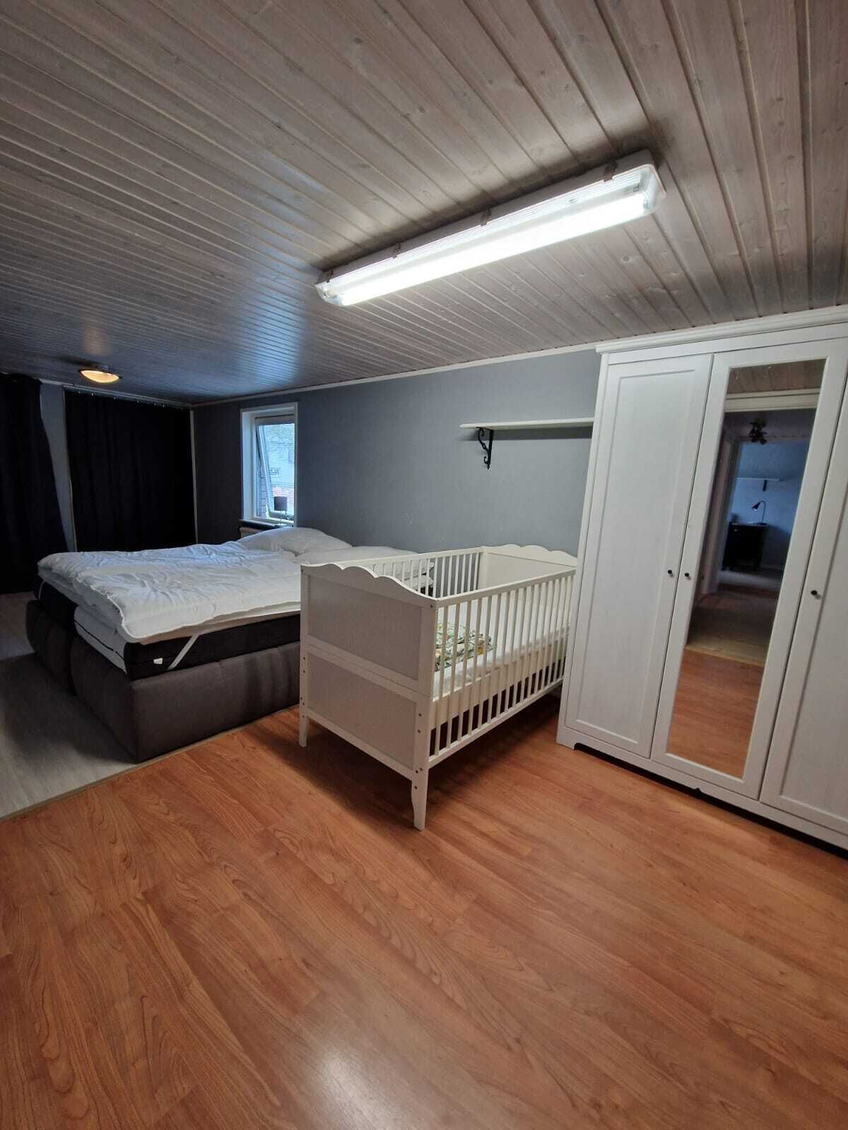 Ein Zimmer mit Holzboden, einem Reisebett, einem Bett, einem Kleiderschrank und einer Deckenleuchte.