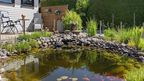 Ein Garten mit Teich, Steinen und Pflanzen. Ein Holzzaun und ein Stuhl sind ebenfalls vorhanden.