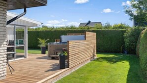 Eine Holzterrasse mit Grill und Sitzgelegenheit, ein Glastür-Eingang und ein gepflegter Rasen.