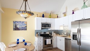 A modern kitchen with white cabinets, stainless steel appliances, a dining table with blue glassware, and a painting on the wall.