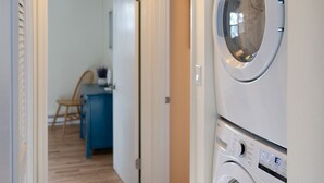 A laundry room with two stacked front-loading washing machines, a wooden floor, and a hallway leading to a dining area.