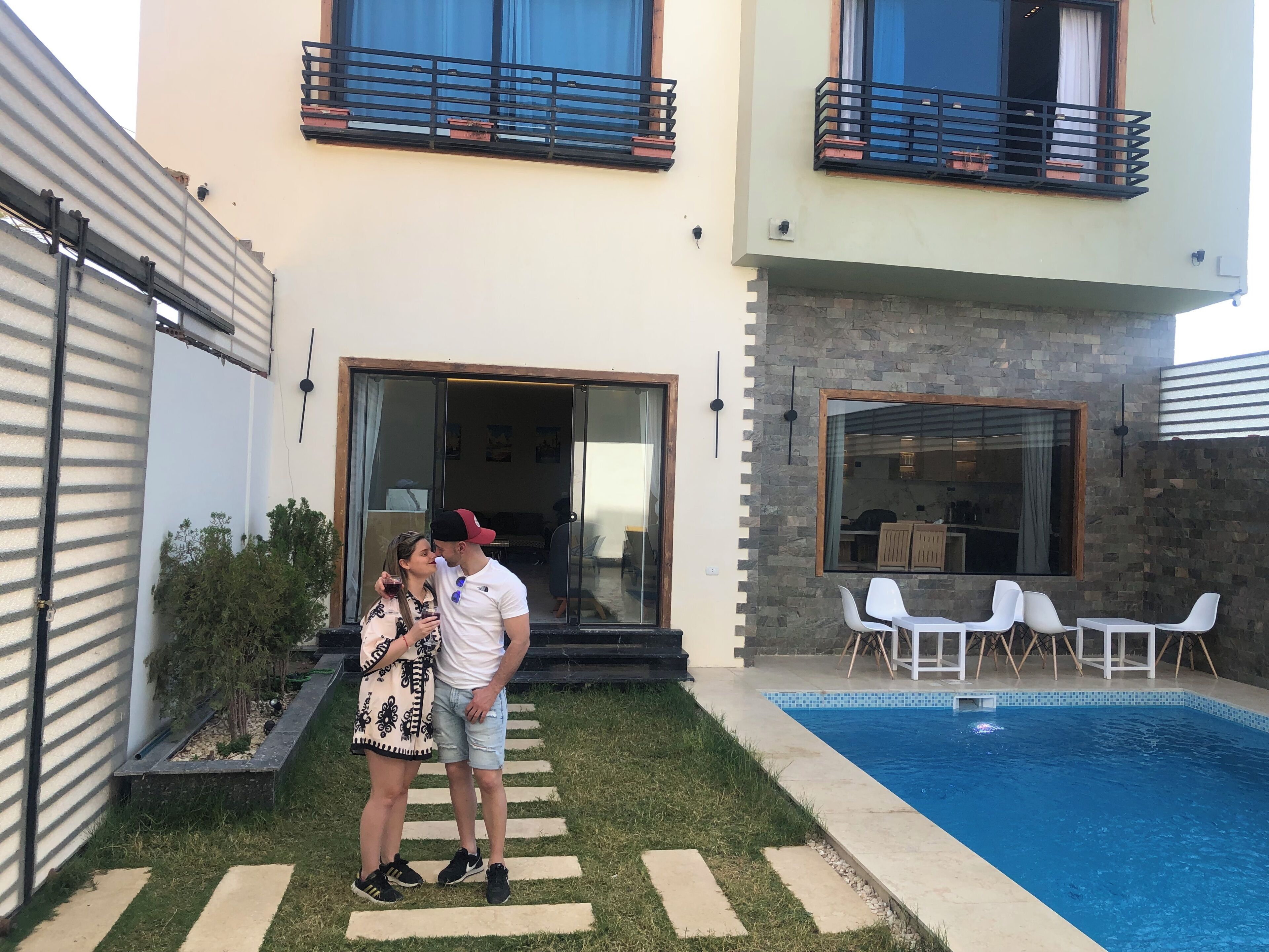 A modern house with a swimming pool, a patio with white chairs, and a glass door leading to a dining area.