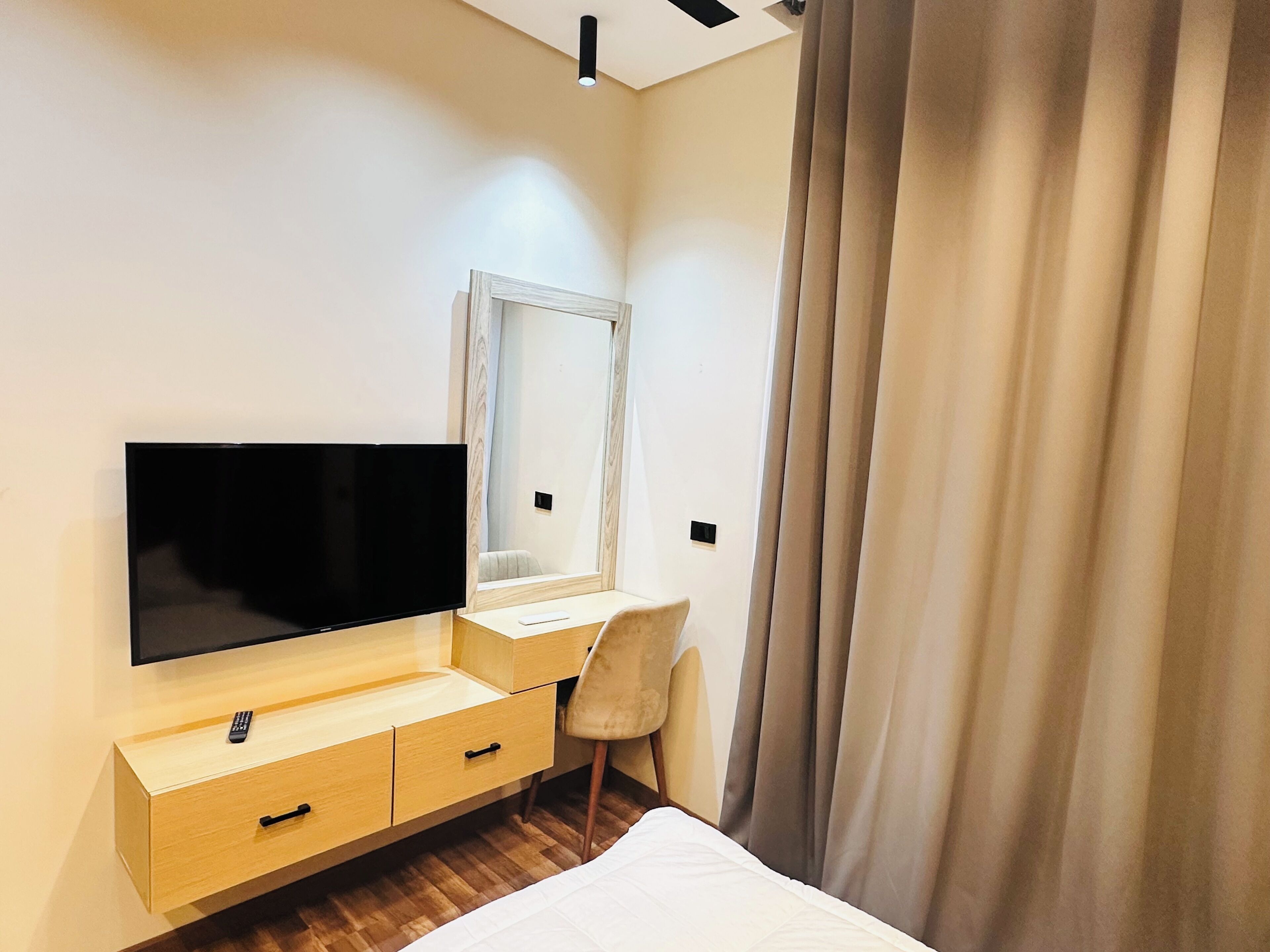 Et moderne hotelværelse med stort fladskærms-tv, træskrivebord, stol, seng og et stort spejl.