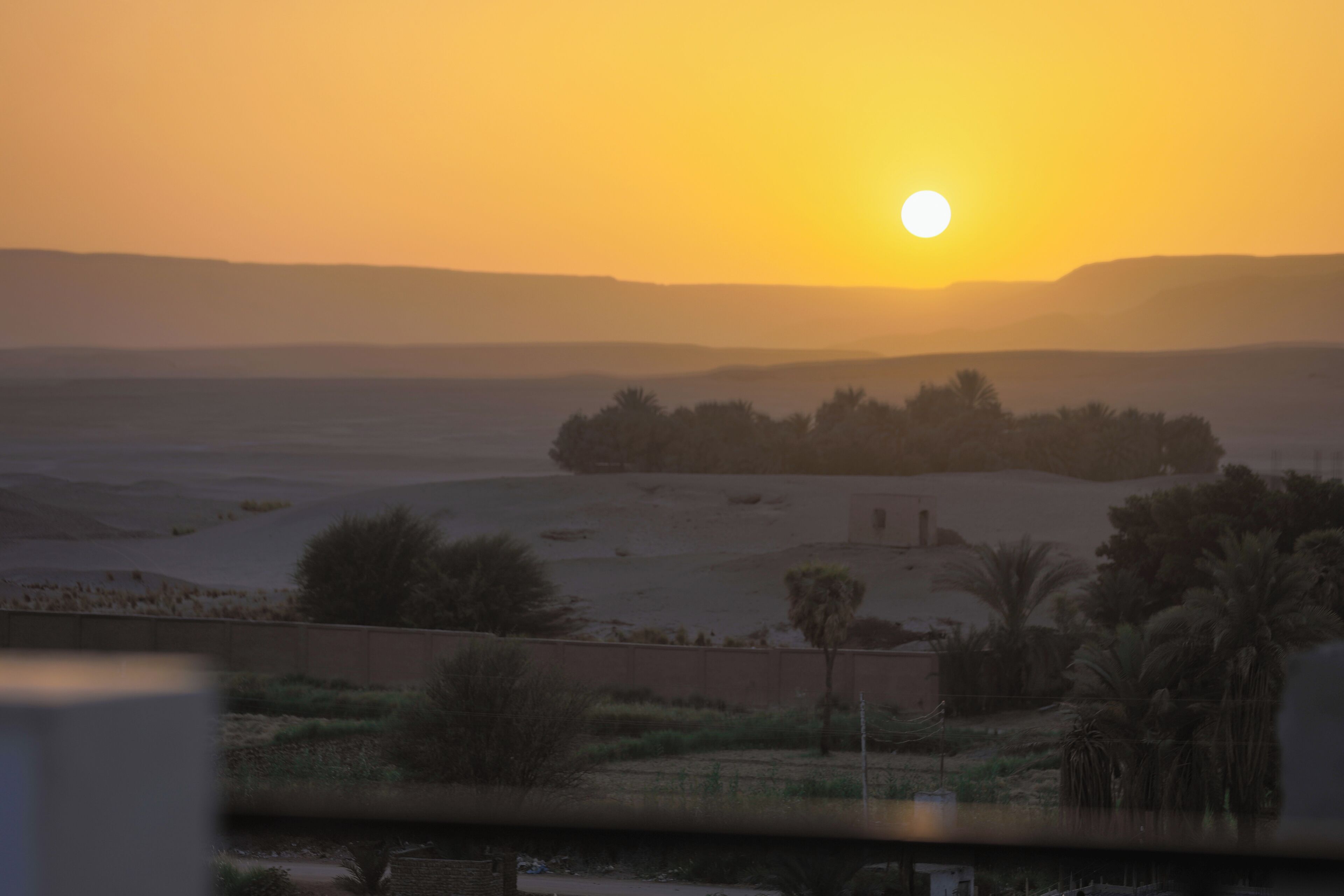 A desert landscape with a sunset, distant hills, and sparse vegetation.