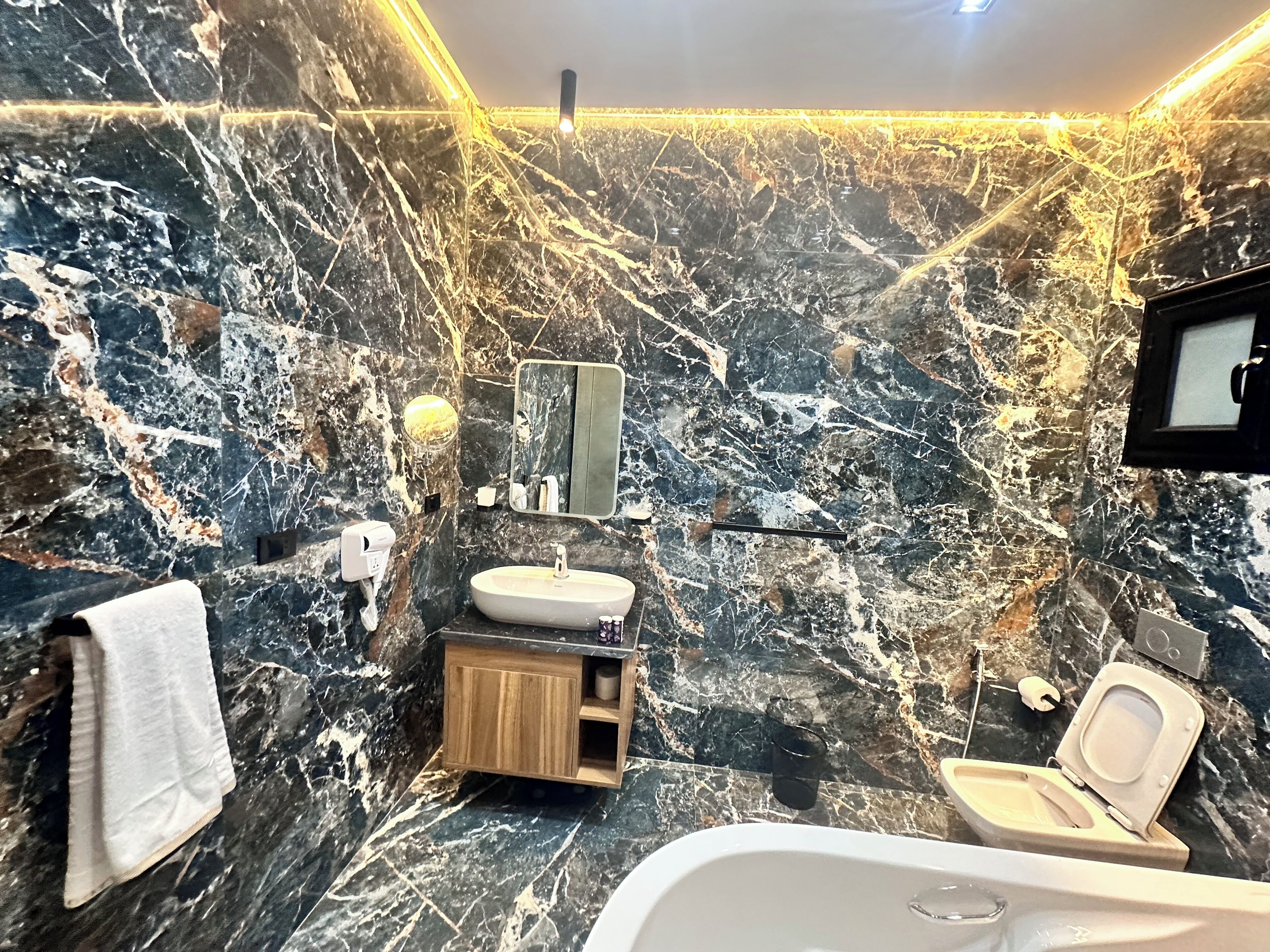 A bathroom with a marble wall, a sink, a mirror, a toilet, and a towel.