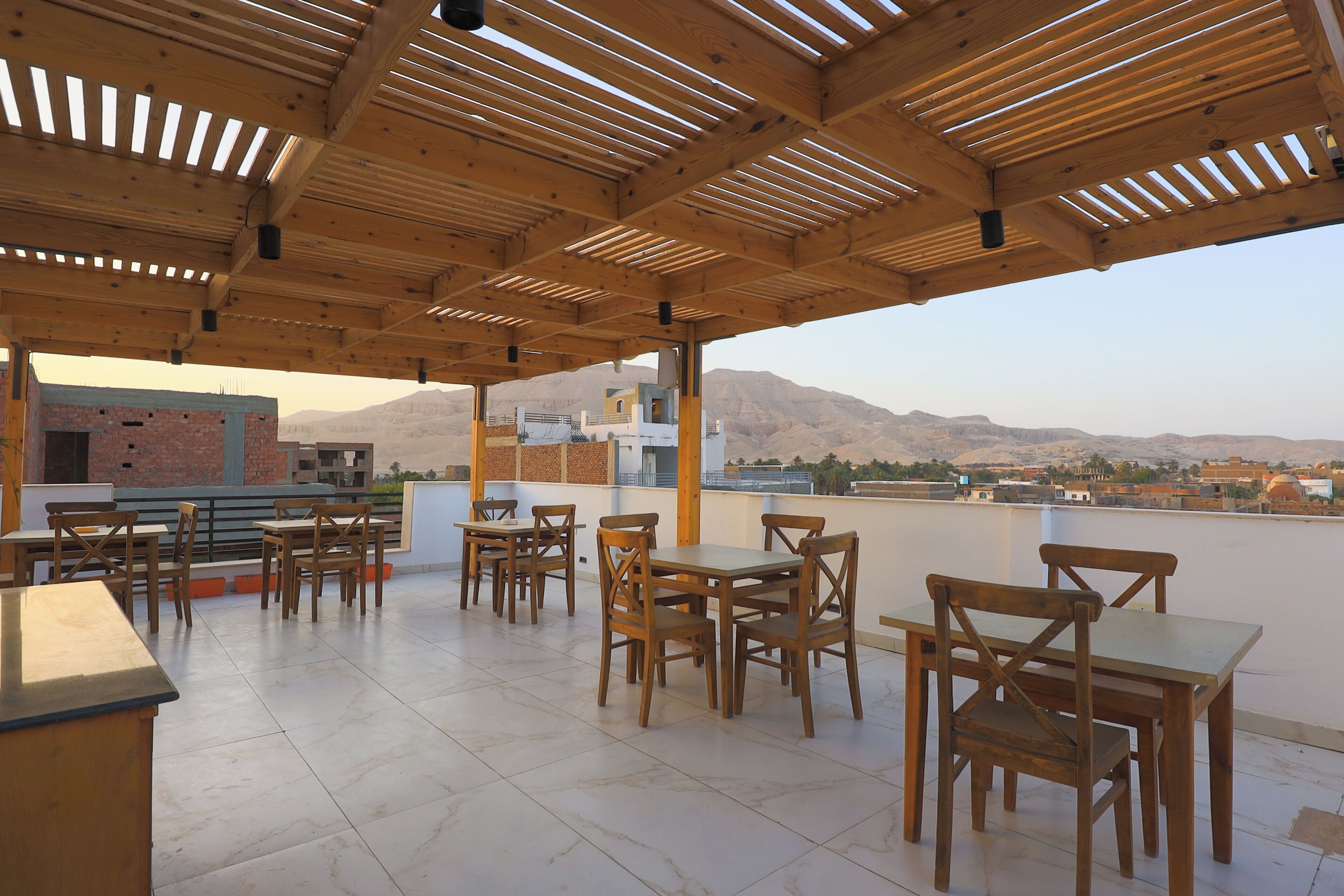 An outdoor patio with wooden tables and chairs, a tiled floor, and a view of a mountainous landscape.