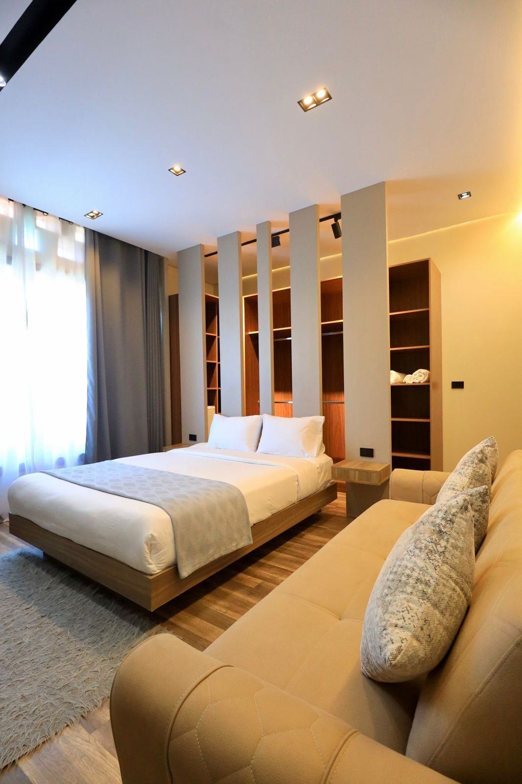 A modern hotel room with a large bed, built-in storage, a sofa, and a large window with curtains.