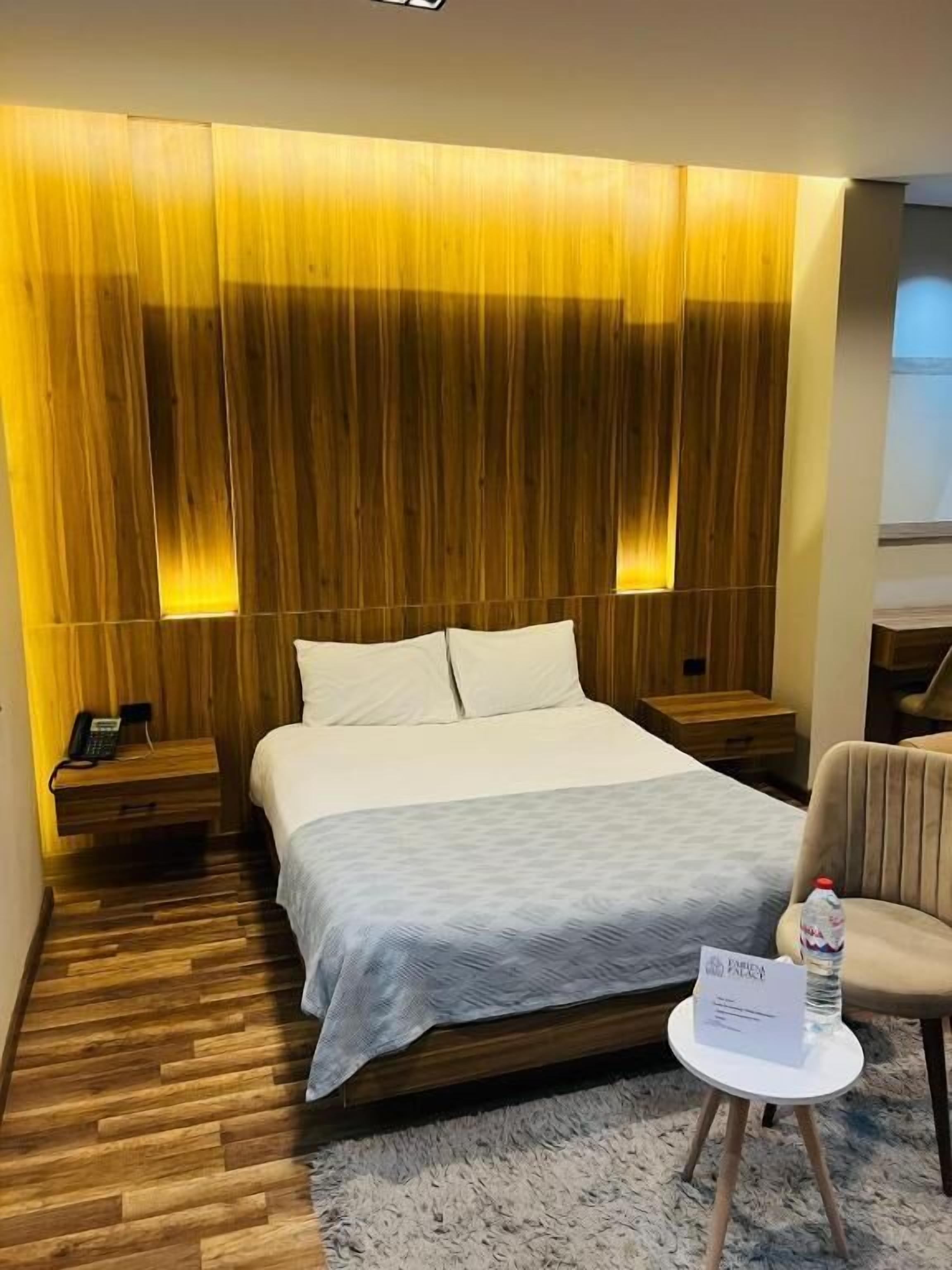 Una camera d'albergo con una testiera in legno, un letto con lenzuola bianche, un comodino con un telefono, un tavolino rotondo con una bottiglia e un cartello, e una sedia.