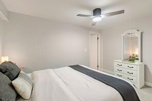 Un dormitorio con una cama, un tocador con cajones, un espejo, un ventilador de techo y una puerta.