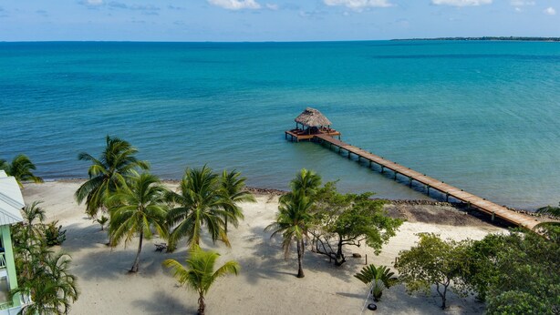 Ein Strand mit einer hölzernen Pier, die zu einem strohgedeckten Gebäude führt, umgeben von Palmen und blauem Himmel.