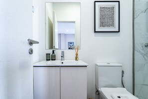 A modern bathroom with a white sink, mirror, and toilet. There is a framed artwork above the sink and a shower with a glass door.