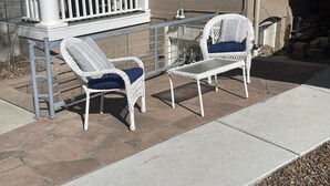 Two white wicker chairs with blue cushions on a patio with a metal railing and a barbecue grill.