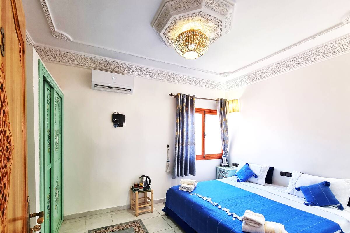 Una habitación de hotel con una cama, una mesita de noche, una puerta verde, una ventana con cortinas y una lámpara decorativa en el techo.