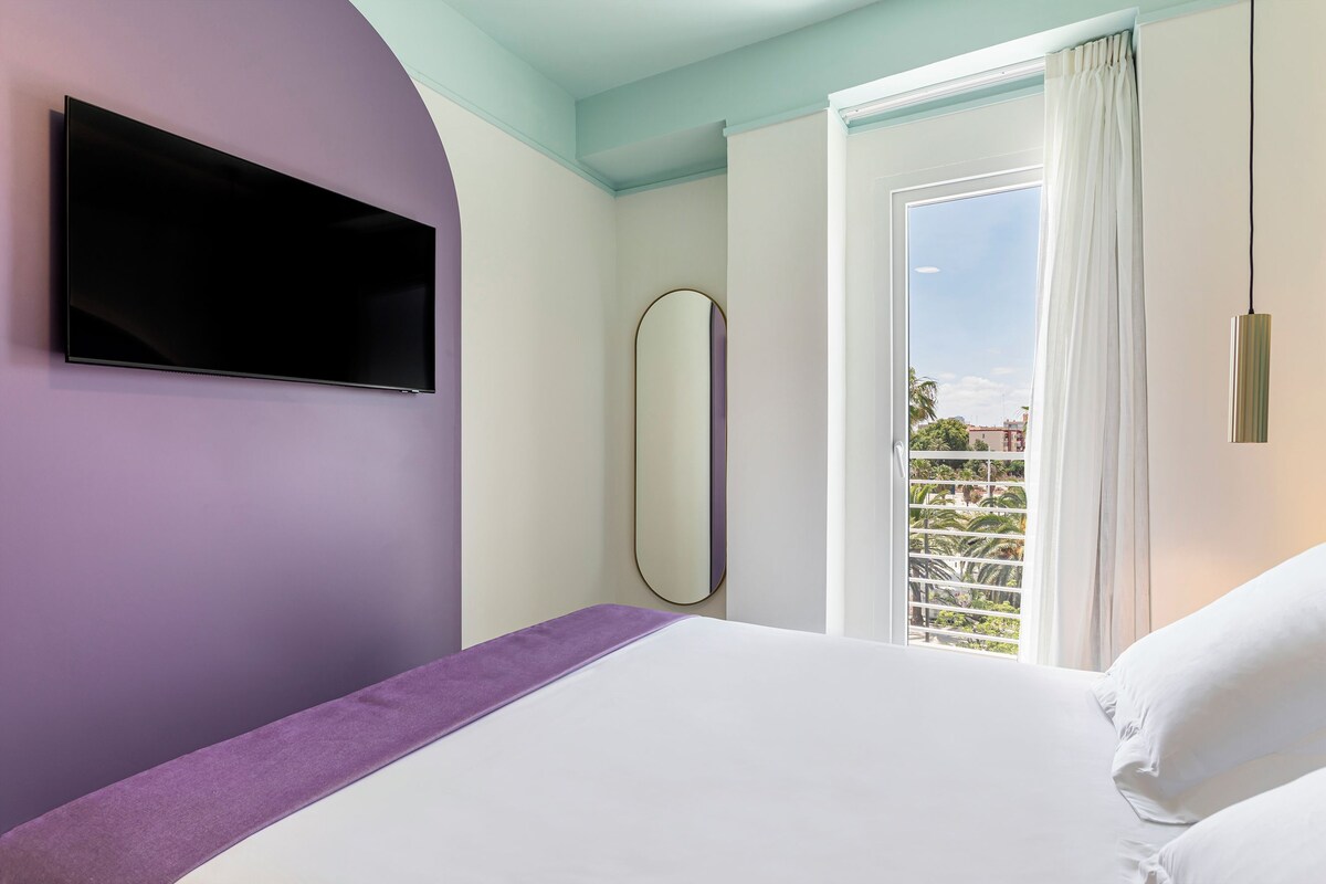 A hotel room with a bed, a flat-screen TV mounted on the wall, a large mirror, and a window with a view of palm trees and buildings.