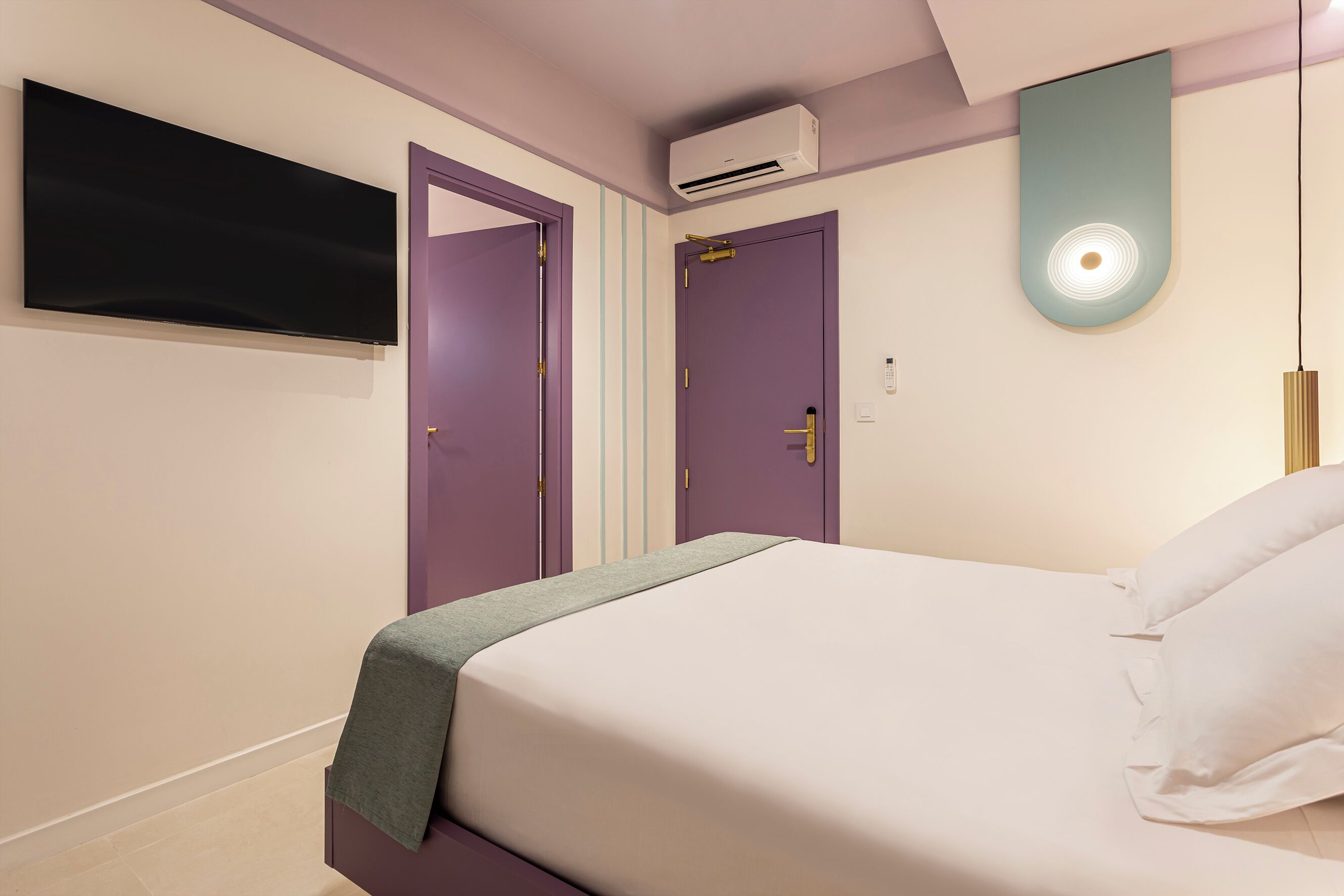 Ein modernes Hotelzimmer mit Bett, Flachbildfernseher, zwei Türen, Klimaanlage und Pendelleuchte.