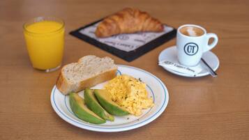 Un plato de desayuno con huevos revueltos, aguacate, tostadas, un croissant, un vaso de jugo de naranja y una taza de café sobre una mesa de madera.