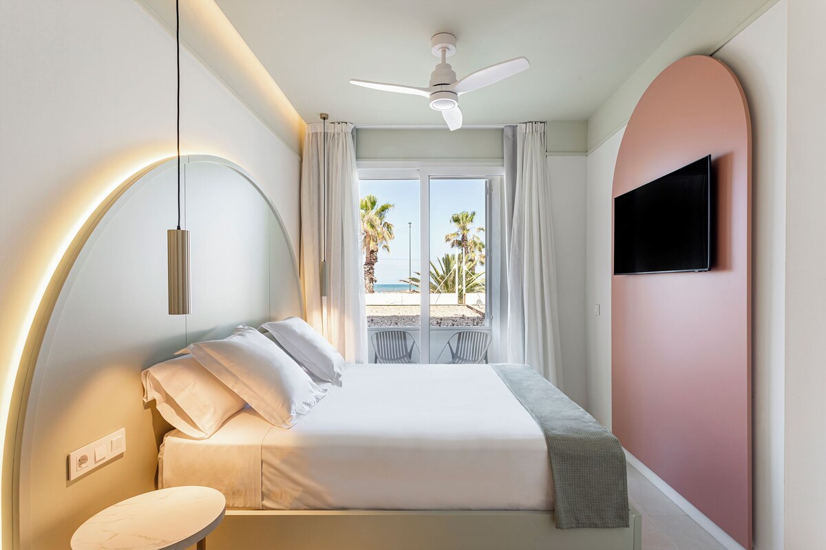 A bedroom with a bed, a ceiling fan, a TV mounted on the wall, and a view of the beach through the window.