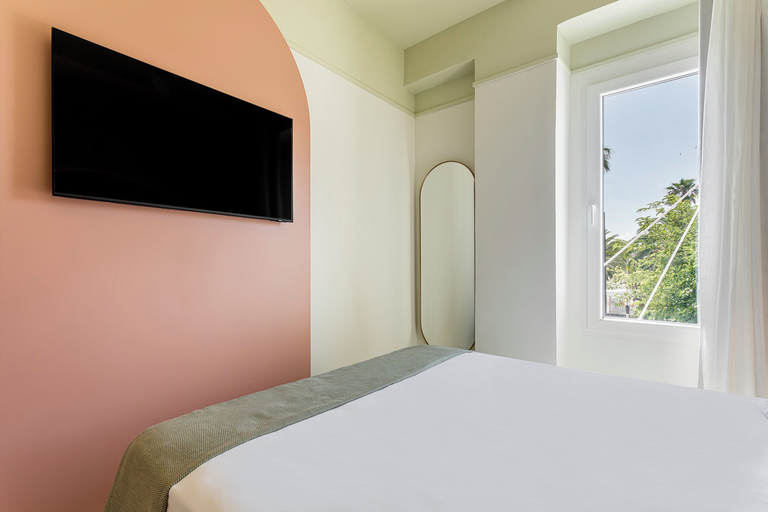 A bedroom with a bed, a TV mounted on the wall, a window with a view of trees, and a door.