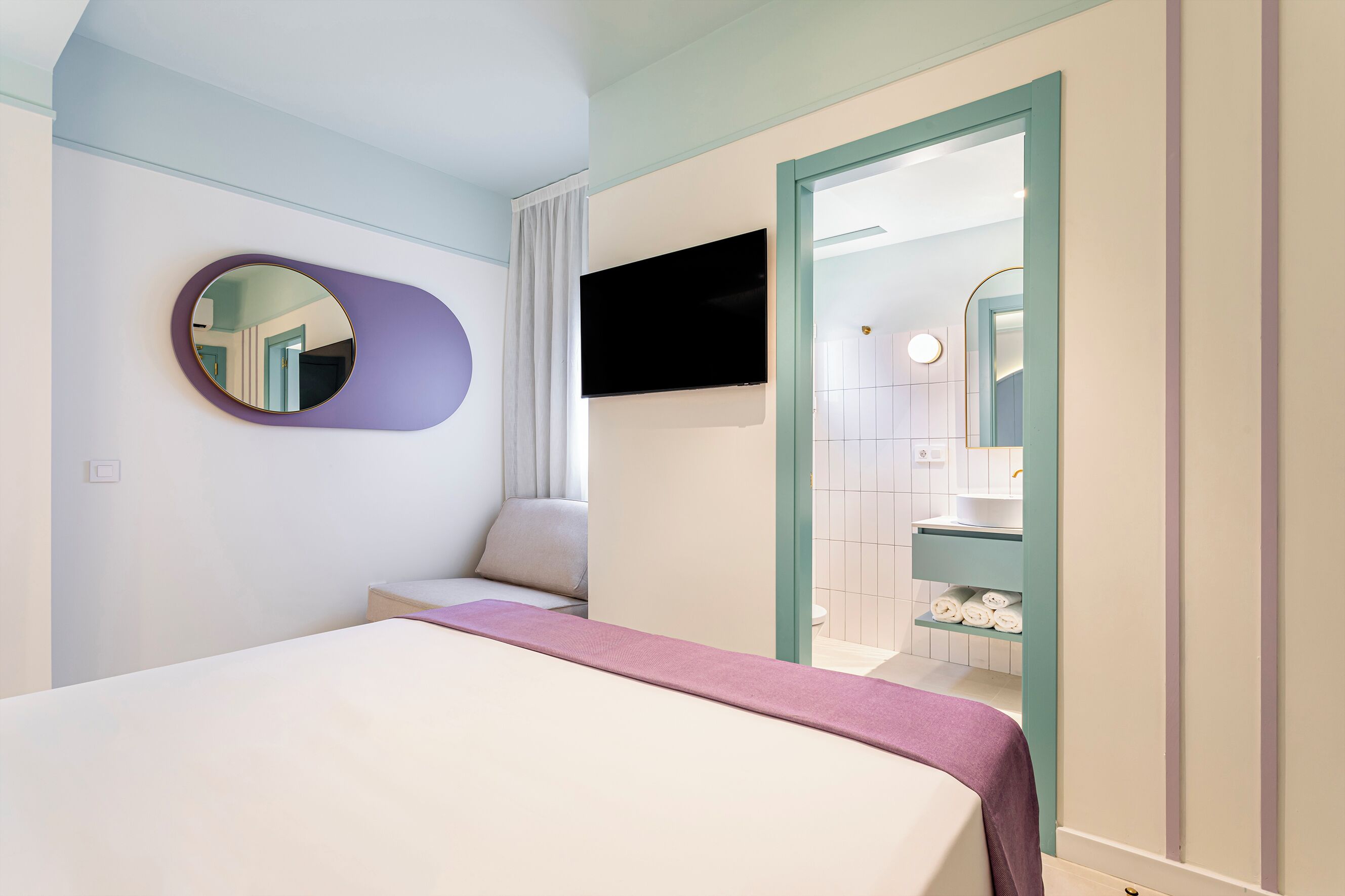 A hotel room with a bed, a round mirror, a television, and a bathroom visible through an open door.
