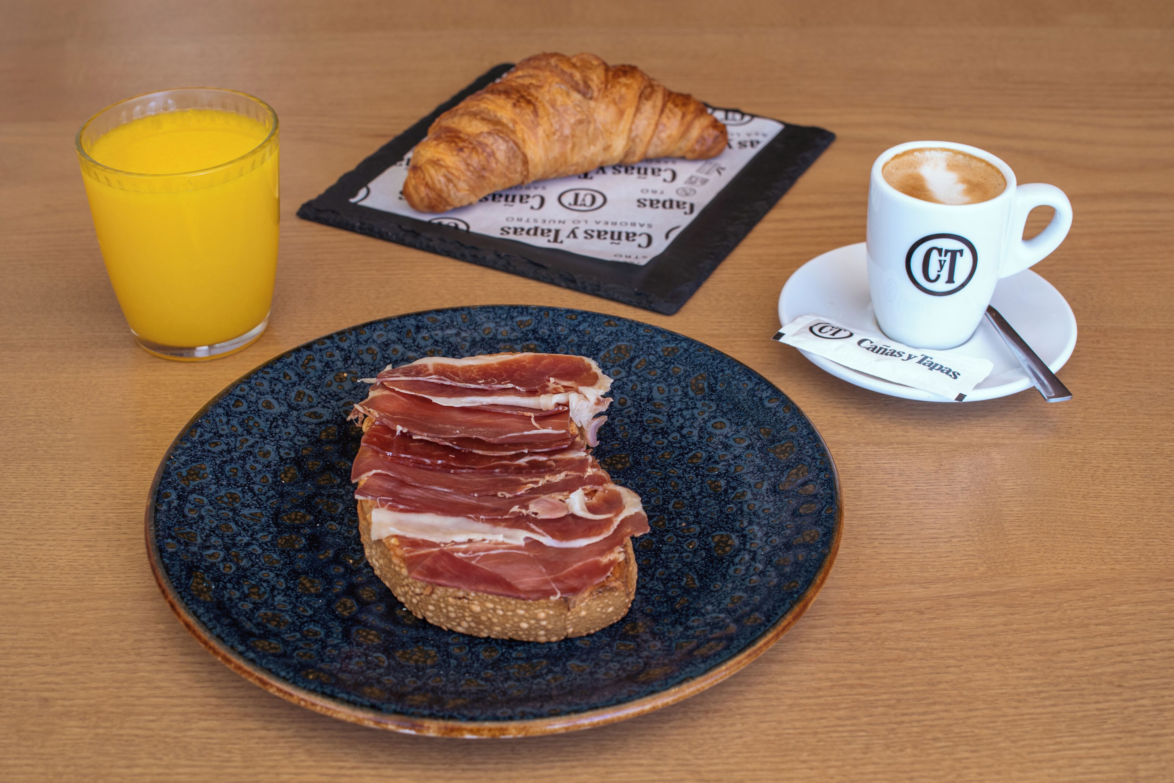 Un desayuno con un croissant, un vaso de zumo de naranja y una taza de café sobre una mesa de madera.
