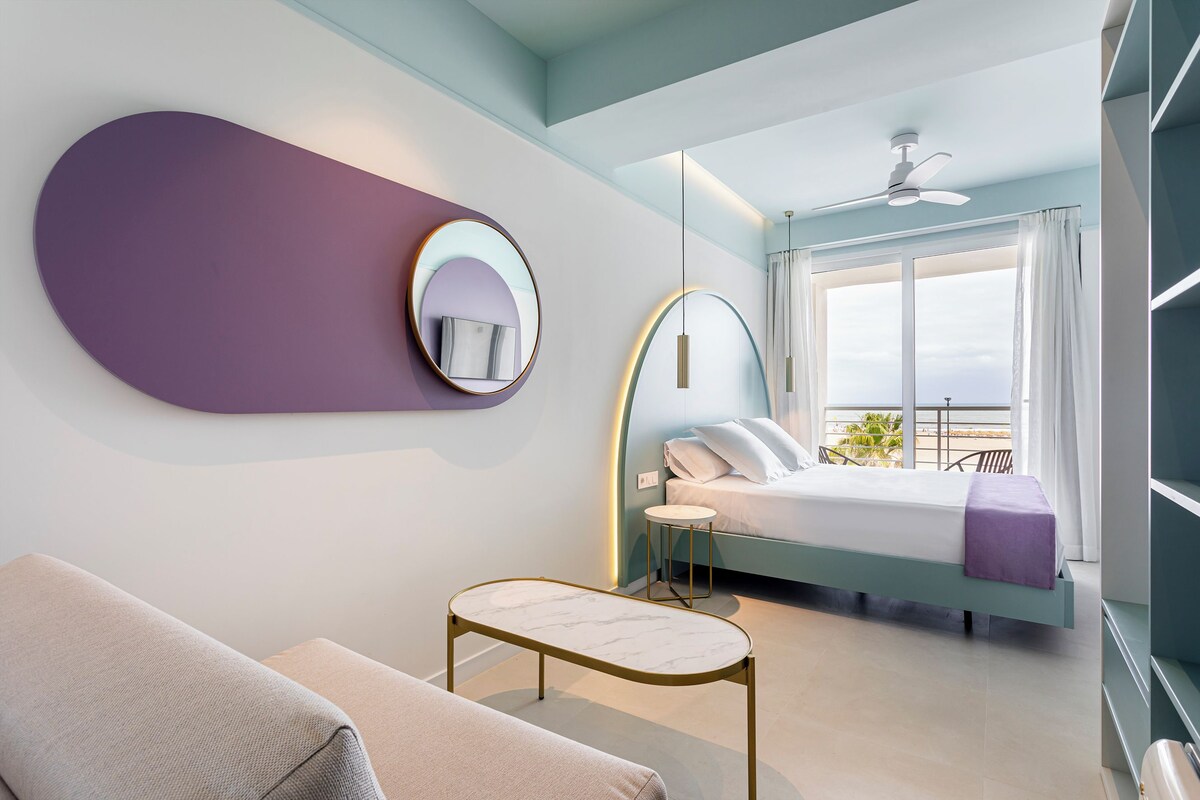 A modern hotel room with a bed, a round mirror, a small table, and a balcony with a view.
