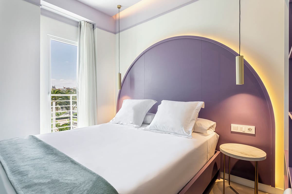 Una habitación de hotel moderna con una cama grande, una lámpara redonda montada en la pared, una mesita redonda y una ventana con cortinas.