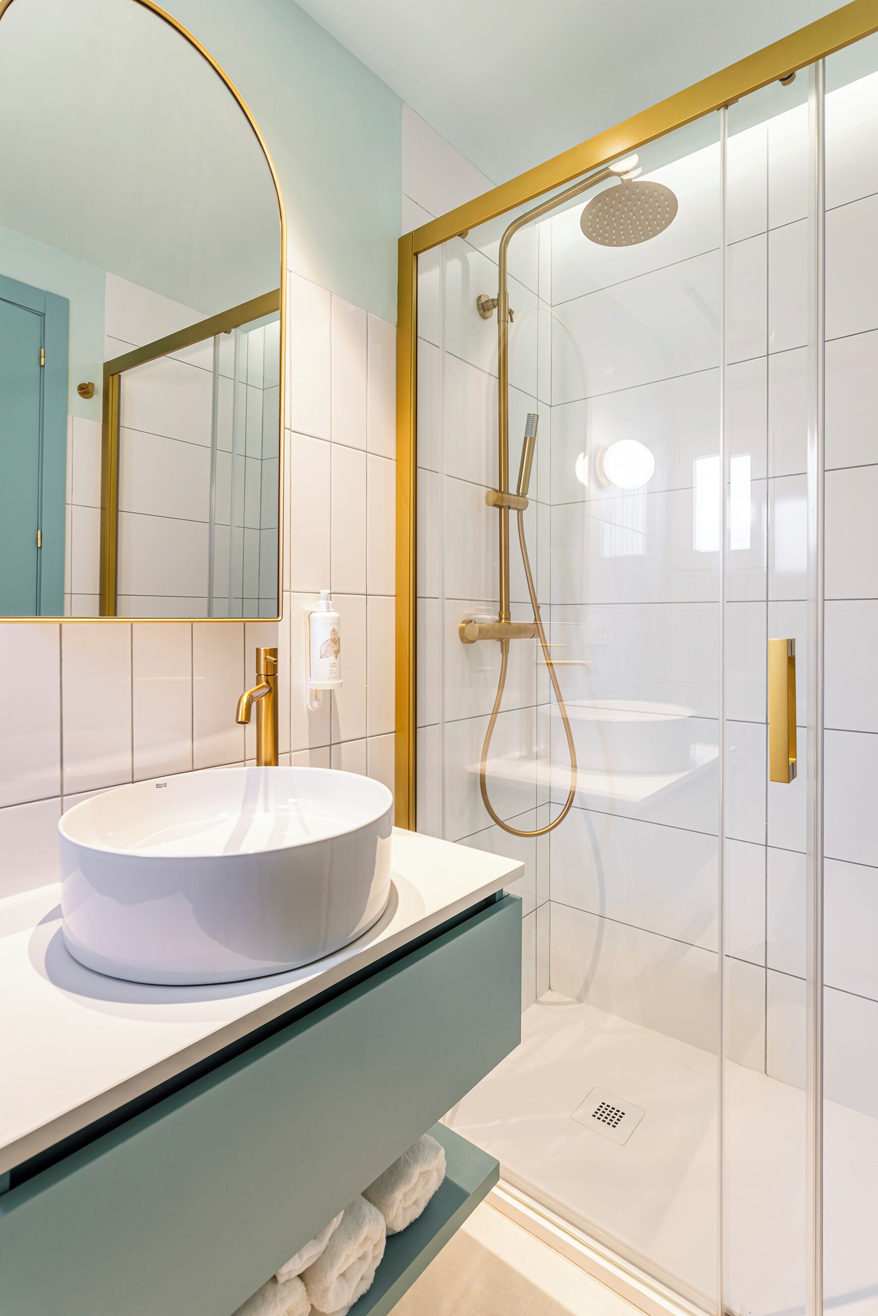 Un baño moderno con un lavamanos blanco redondo, un grifo dorado, ducha con mampara de vidrio y un espejo con marco dorado.
