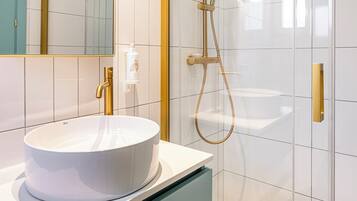 Un baño moderno con un lavamanos blanco redondo, un grifo dorado, ducha con mampara de vidrio y un espejo con marco dorado.
