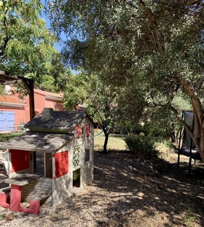 A small playhouse with a red roof and white walls, surrounded by trees and a gravel path.