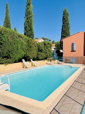 Ein rechteckiger Pool mit einer gefliesten Kante, umgeben von einer niedrigen Mauer. Zwei Liegestühle und ein kleiner Tisch mit einem blauen Sonnenschirm stehen bereit.