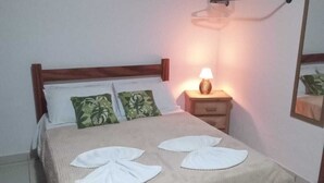 Cama bem arrumada com colcha bege e duas toalhas decorativas, abajur e ar condicionado fixado na parede.