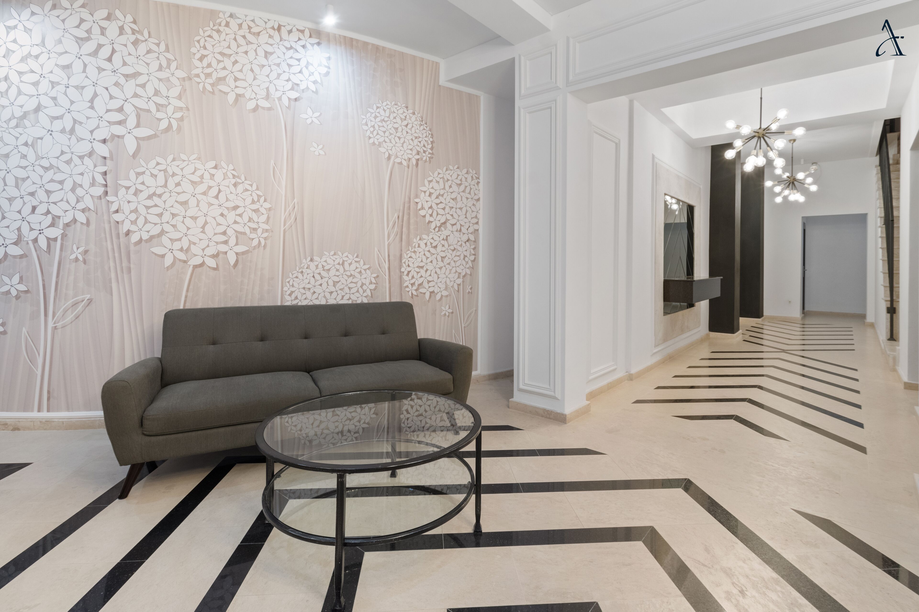A modern living room with a sofa and a glass coffee table, a decorative wall with floral patterns, and a geometric patterned floor.