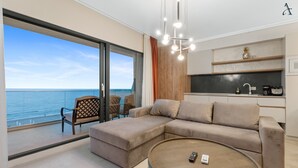 A modern living room with a large window overlooking the sea, a beige sectional sofa, a round coffee table, and a minimalist kitchen area. - Gioia Sea View (Navodari)