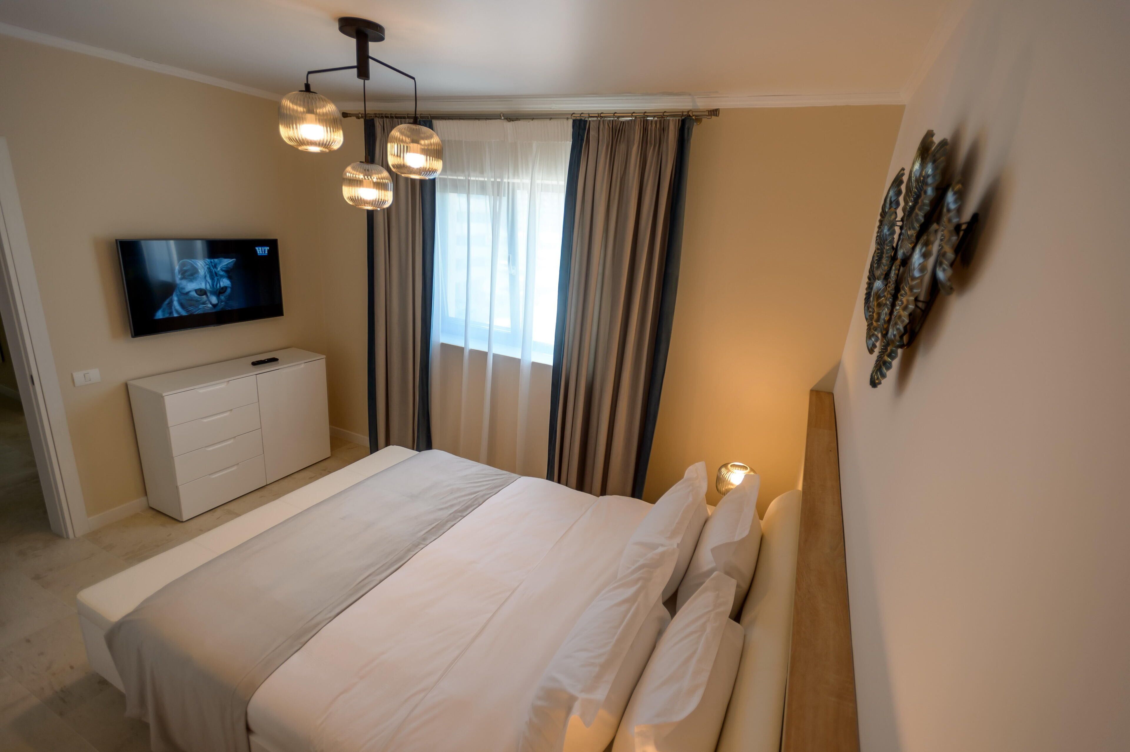 A bedroom with a bed, a TV mounted on the wall, a dresser, and a window with curtains.