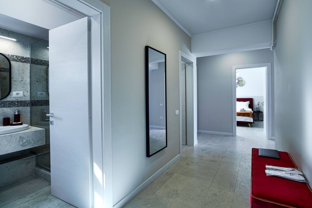 A modern hotel hallway with a large mirror, a red bench, and a bed in the background.