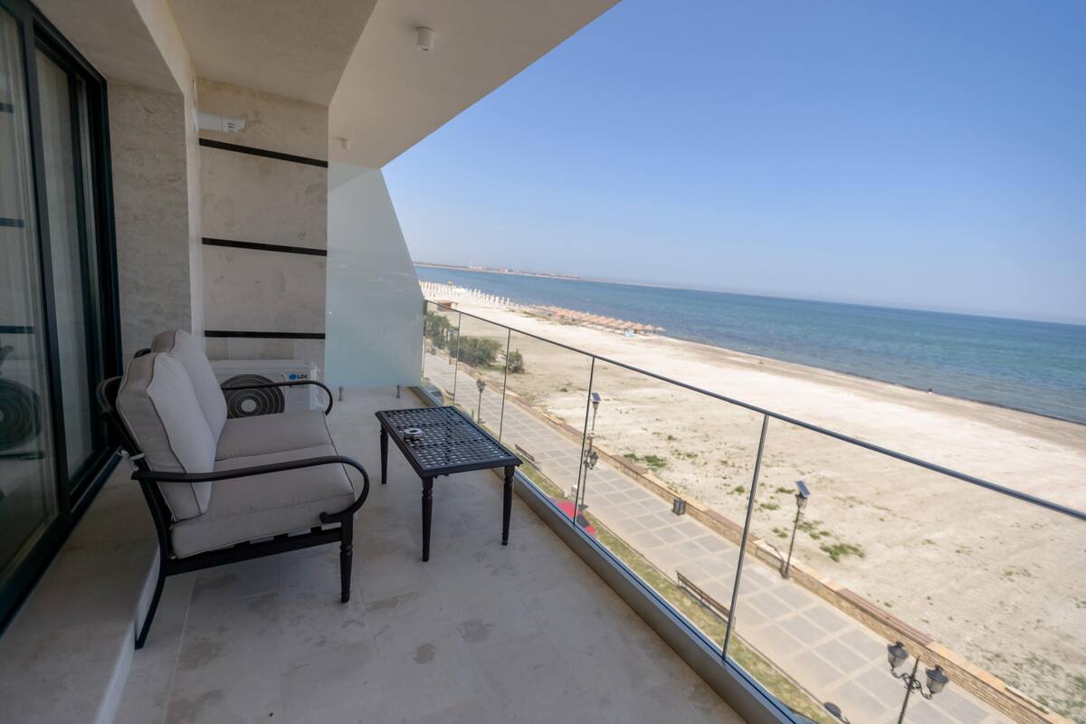 A balcony with a view of the beach, a chair, a small table, and a glass door.
