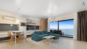 A modern living room with a teal sofa, a white coffee table, and a dining area with a white table and chairs. A kitchen with white cabinets and a microwave is visible. The room has a large window with a view of the sea. - Gioia Sea View (Navodari)