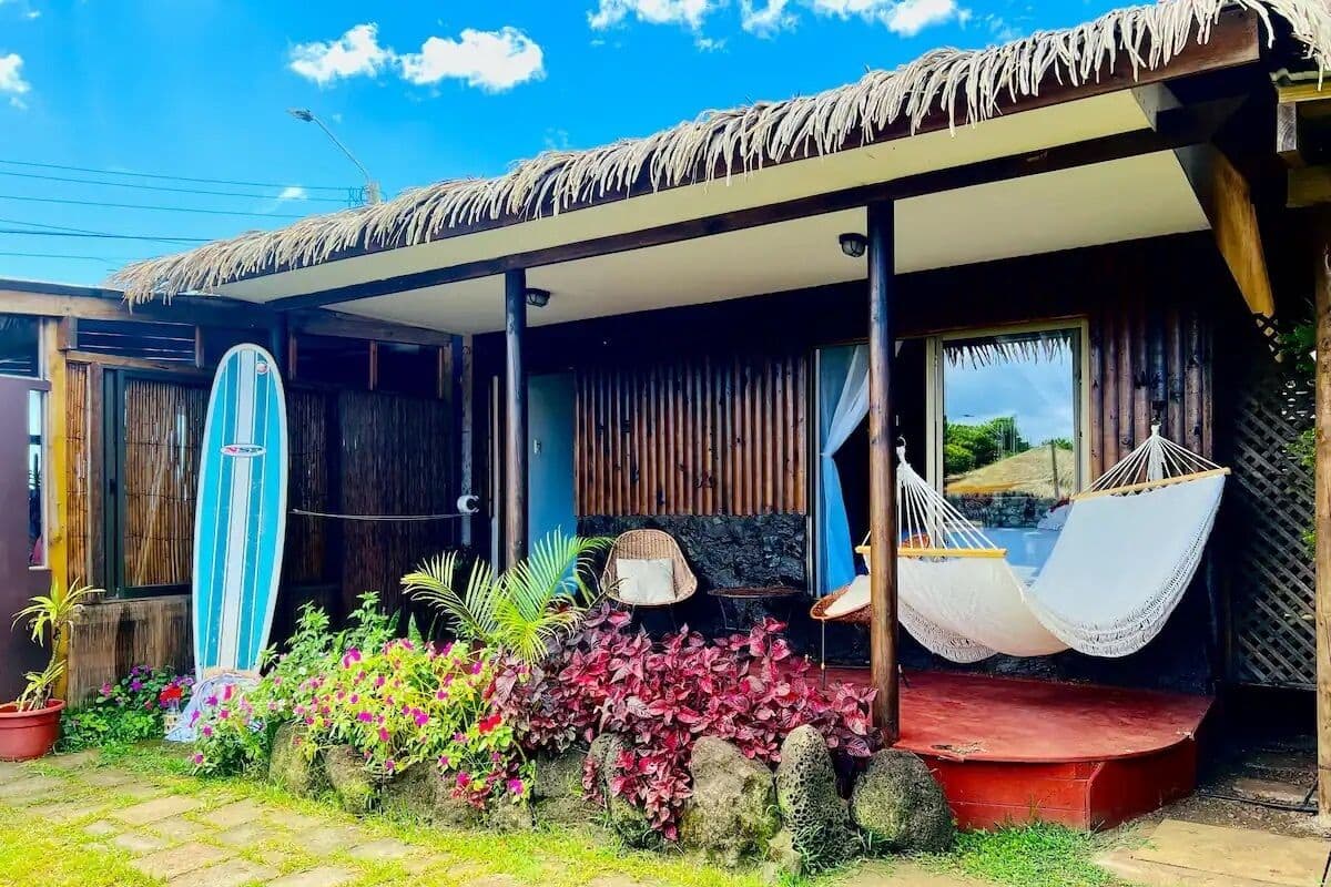 A beach house with a thatched roof, a surfboard leaning against the wall, and a hammock on the porch.